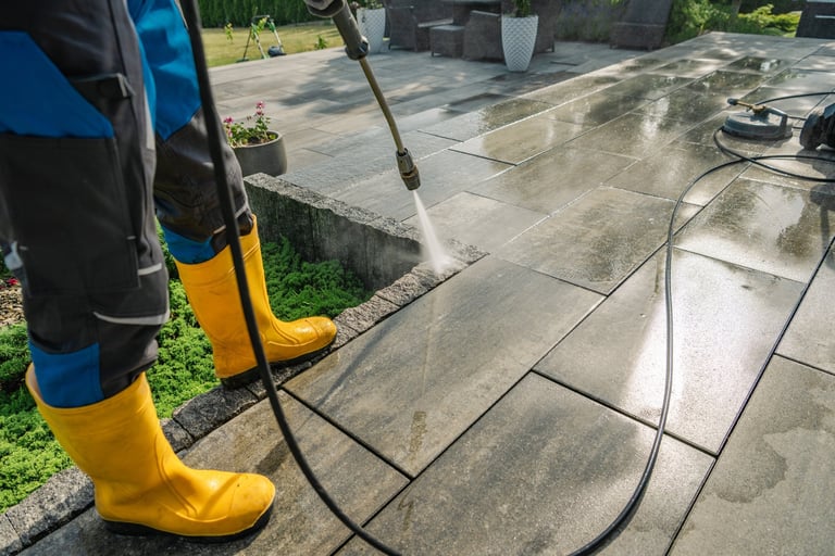 A worker in yellow boots cleans a stone patio with a pressure washer on a sunny day
