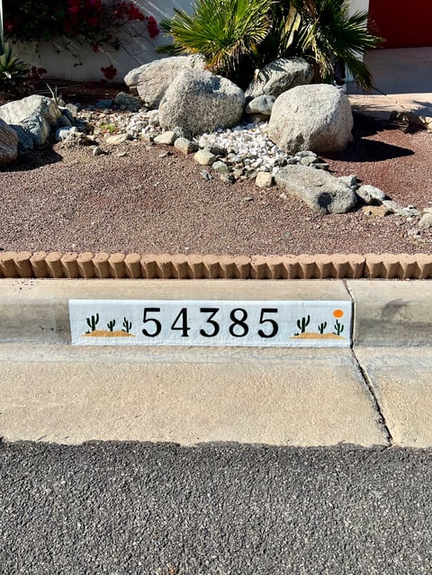 Desert landscaping with large boulders, white rocks, and a palm tree behind a house address marker reading 54385