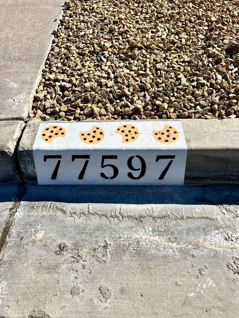 White marker labeled 77597 with four chocolate chip cookies on top, placed on gravel and concrete at an archaeological or construction site