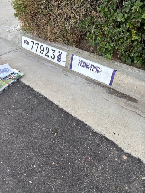 White sign displaying number 77923 and text on concrete curb next to asphalt and overgrown vegetation