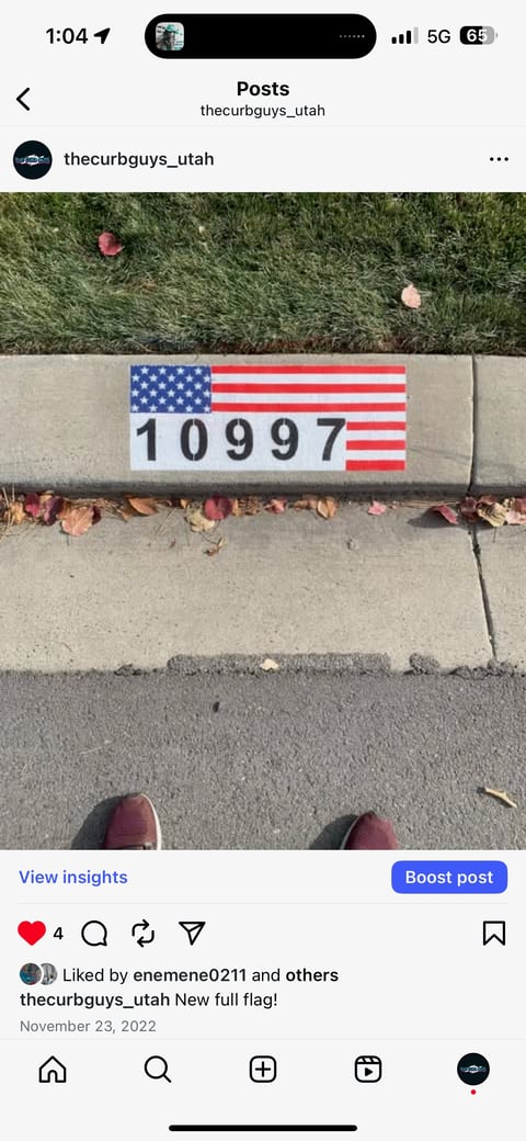 American flag painted on curb with number 10997, two peoples feet visible at bottom, grass and fallen leaves above