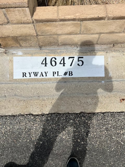 White address label on brick wall showing 46475 Ryway Pl #B with shadow of person taking photo