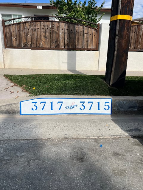 Blue and white address marker displaying 3717-3715 on driveway in front of residential property with wooden gate
