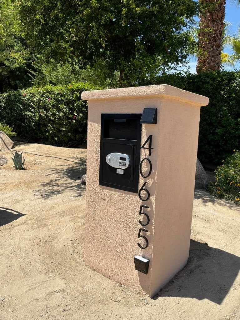 Beige stucco mailbox with black door and address numbers 40655 in a residential garden setting with green foliage