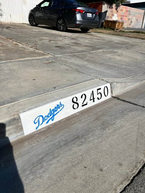 White parking space marker with Dodgers logo and number 82450 on concrete lot with dark sedan in background